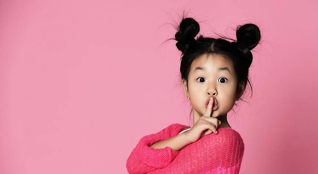 A cute young girl is wearing a pink jumper against a pink background and holding her finger to her lips.