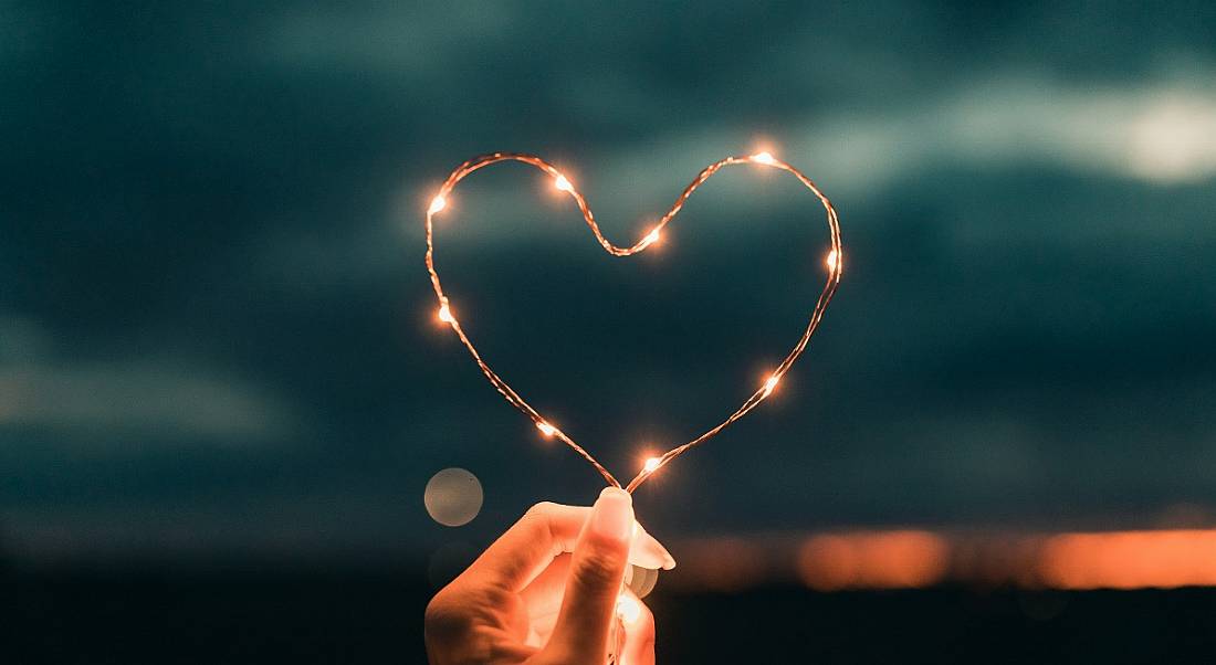 A person's hand is holding fairy lights in the shape of a heart at night.