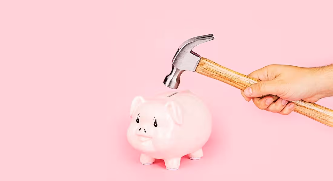 A hand is holding a hammer close to a pink piggy bank against a pink background.