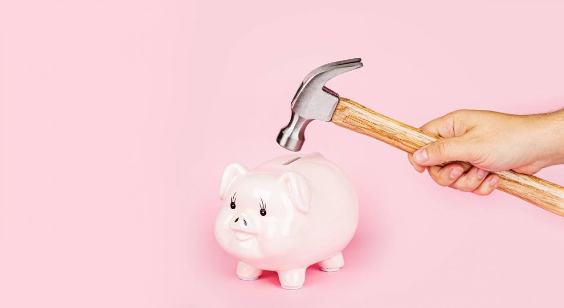 A hand is holding a hammer close to a pink piggy bank against a pink background.