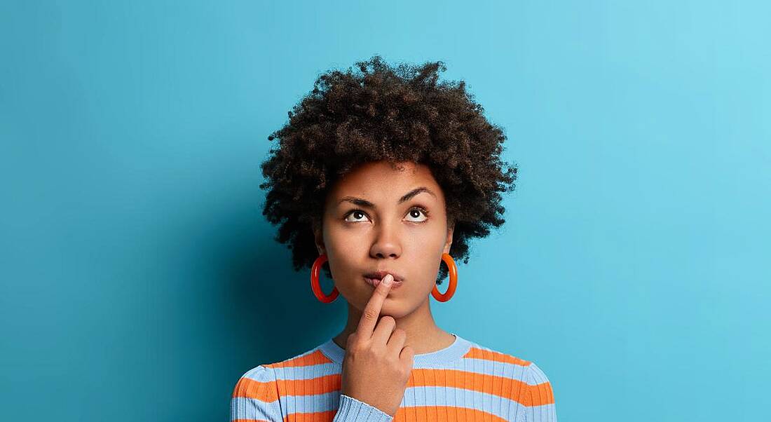 A woman is looking up with her finger against her lips as if making a decision. She is wearing an orange and blue striped jumper against a blue background.