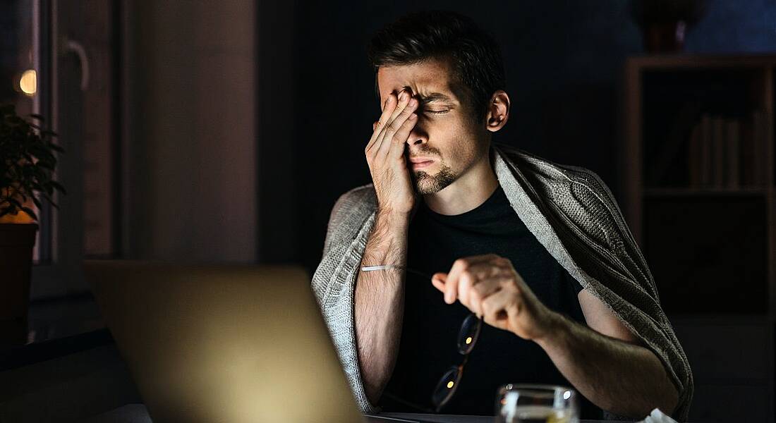Exhausted man rubbing his eyes while looking at his laptop in a dark room.