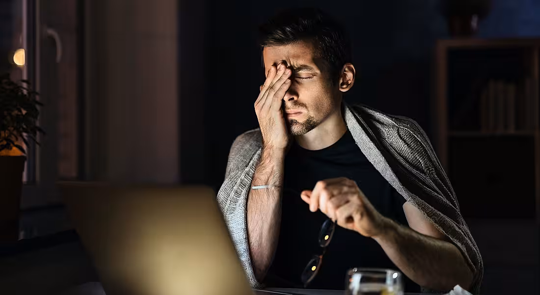 Exhausted man rubbing his eyes while looking at his laptop in a dark room.