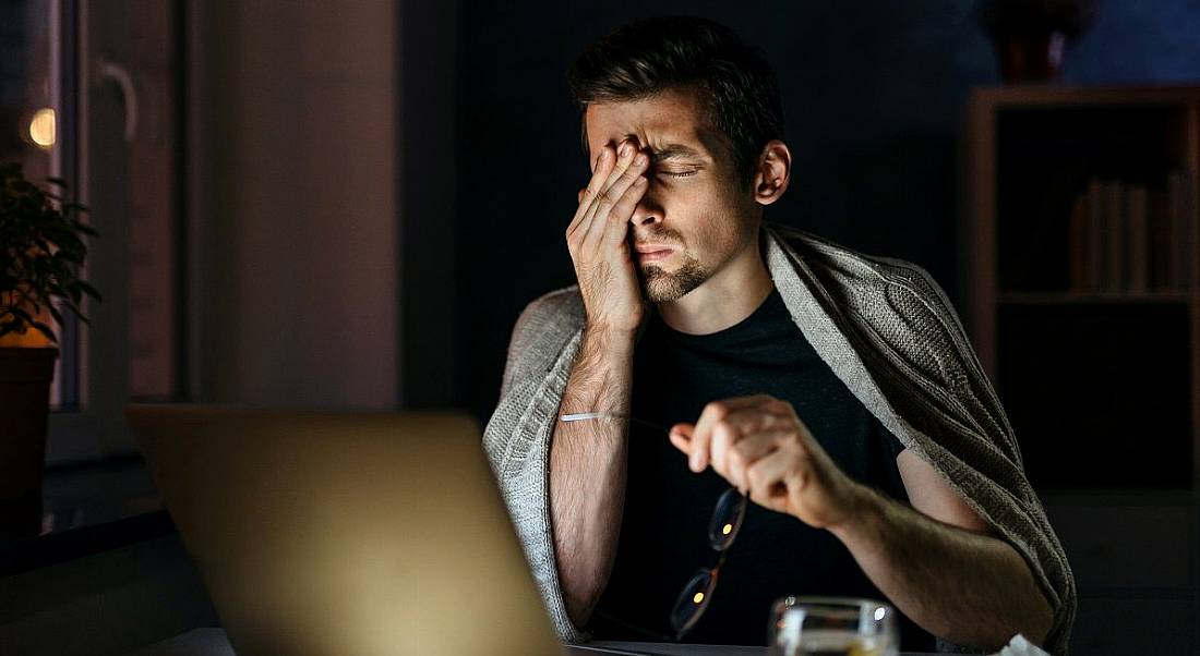 Exhausted man rubbing his eyes while looking at his laptop in a dark room.