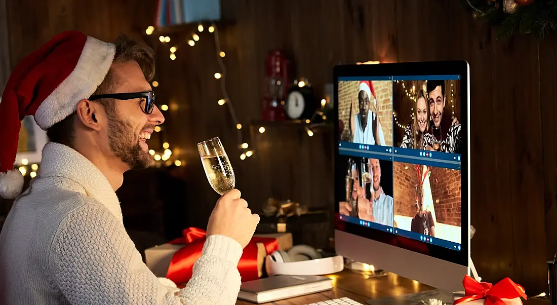 A man is drinking champagne and talking to colleagues on a video call at a virtual Christmas party.