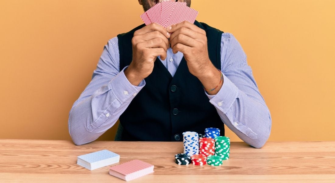 Cropped image of a man holding up playing cards at a desk with poker chips and two piles of playing cards.