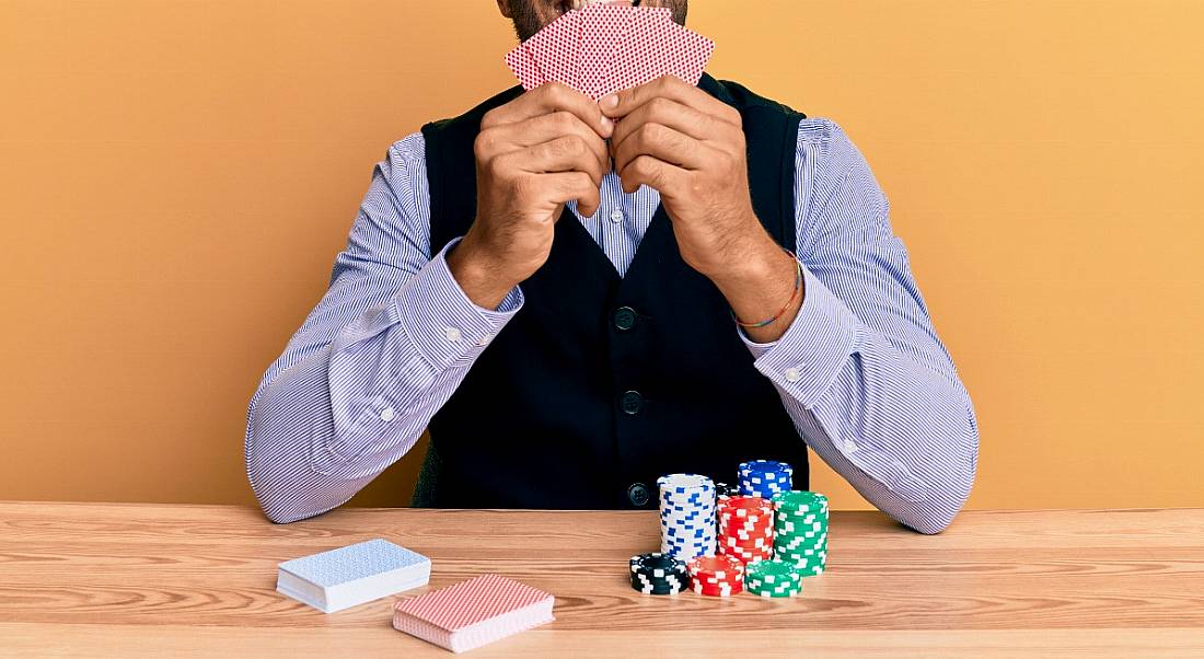 Cropped image of a man holding up playing cards at a desk with poker chips and two piles of playing cards.