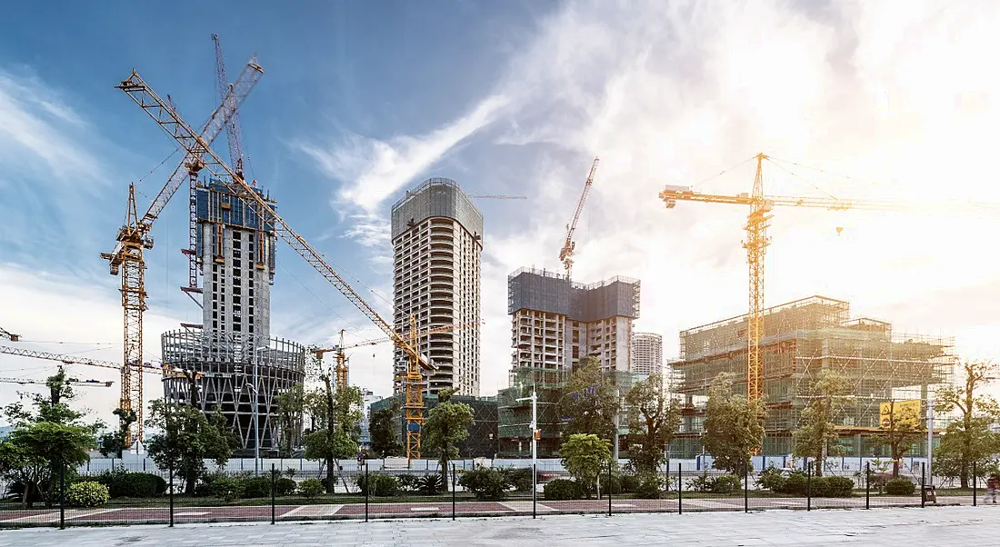 A construction site building a campus of large, tall buildings, with numerous yellow cranes in operation, next to a tree-lined street.