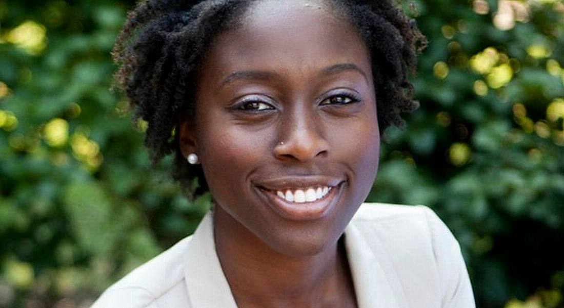 Jamie Adasi of Greenhouse is sitting outdoors in front of foliage, smiling into the camera while wearing a white top.