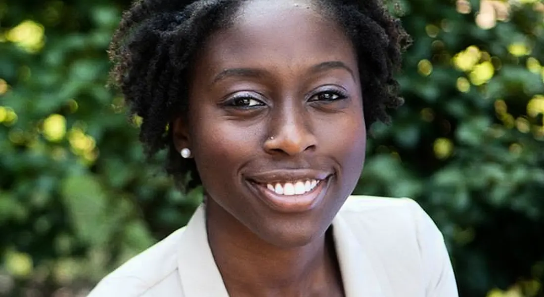 Jamie Adasi of Greenhouse is sitting outdoors in front of foliage, smiling into the camera while wearing a white top.