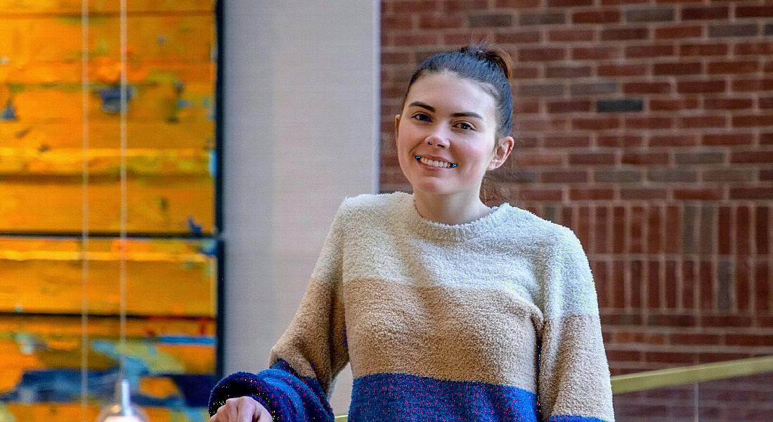 Catie Pitts, HR business partner at Genuity Science, is standing in front of a red brick wall in an office while smiling into the camera. She is wearing a striped jumper and resting her arm on a handrail.