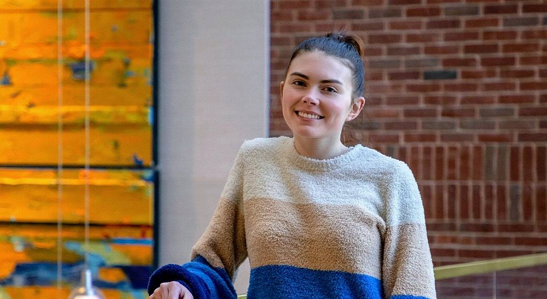 Catie Pitts, HR business partner at Genuity Science, is standing in front of a red brick wall in an office while smiling into the camera. She is wearing a striped jumper and resting her arm on a handrail.