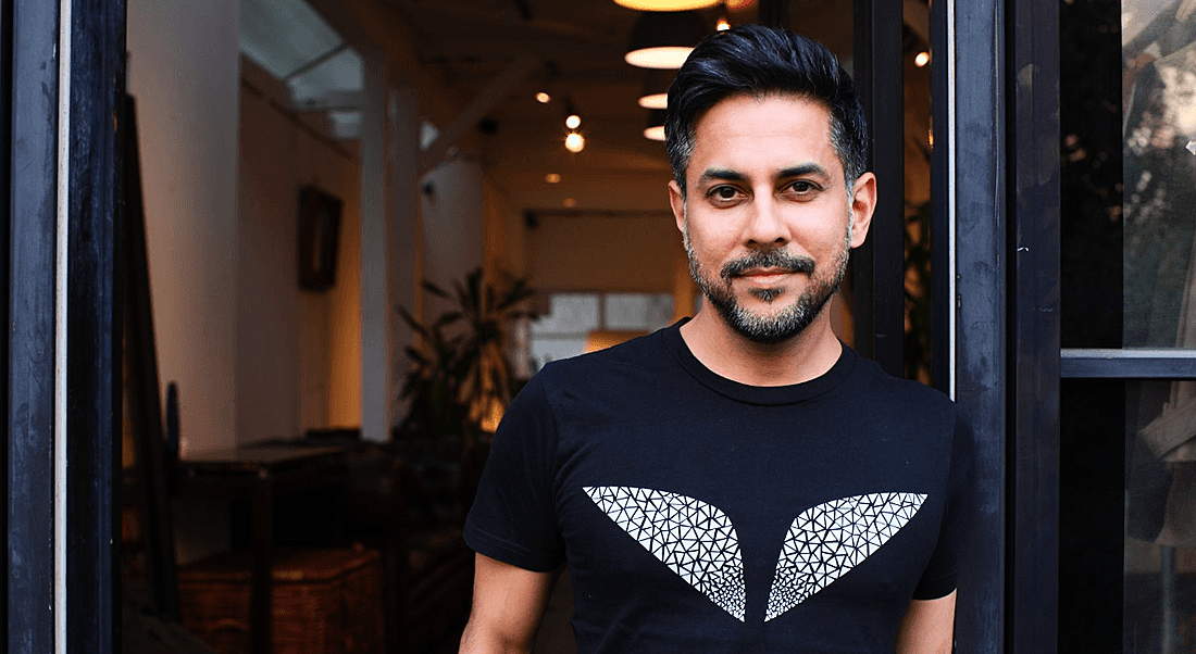 Vishen Lakhiani of Mindvalley is standing in the doorway of a dimly lit room and smiling into the camera.