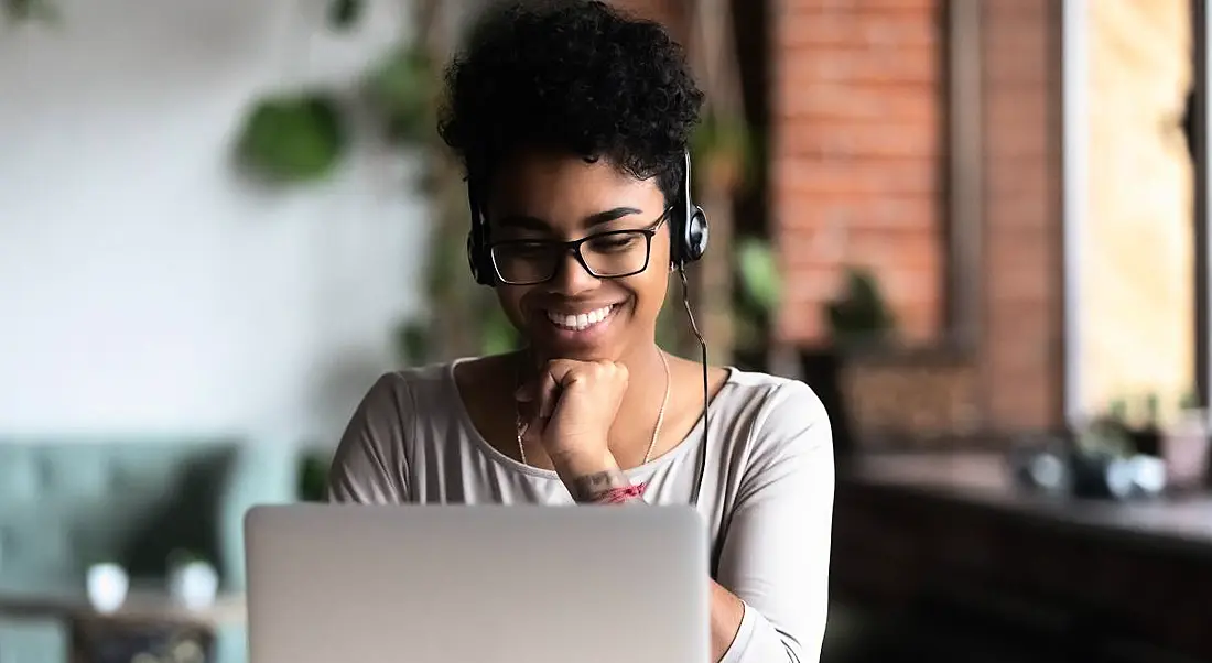 A woman college student smiling while looking at her laptop. She’s wearing headphones and glasses.