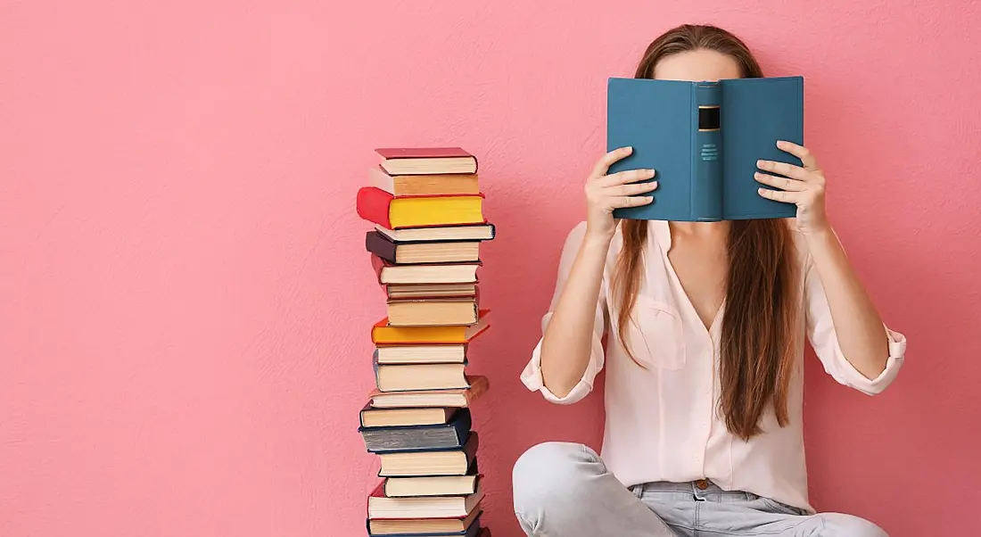 Woman sitting on a floor and reading a book against a pink background, symbolising going back to school during your career.