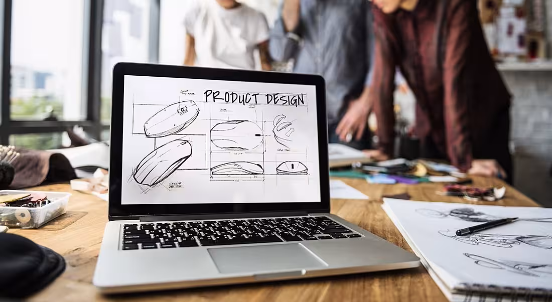 A team of product designers and developers are in the background discussing a project with a laptop in the foreground showing product design.
