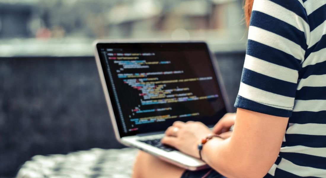 A young woman is coding on a laptop outdoors while wearing a black and white striped t-shirt.