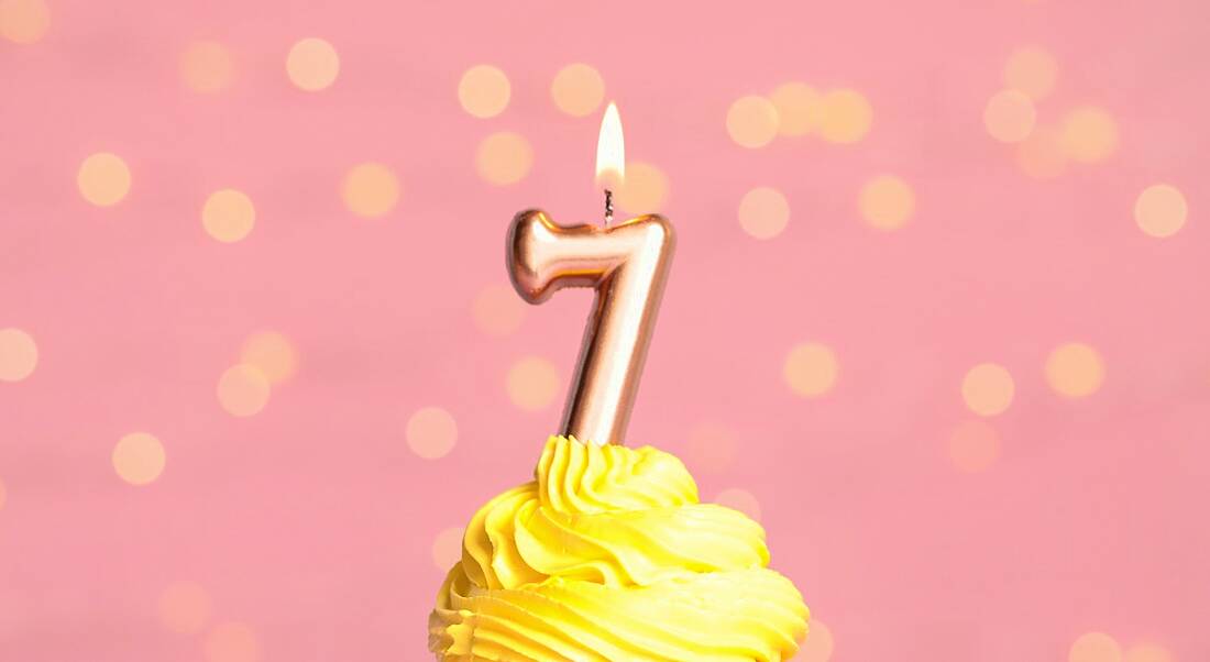 Birthday cupcake with yellow icing and a number seven birthday candle on top, sitting against a pink background.