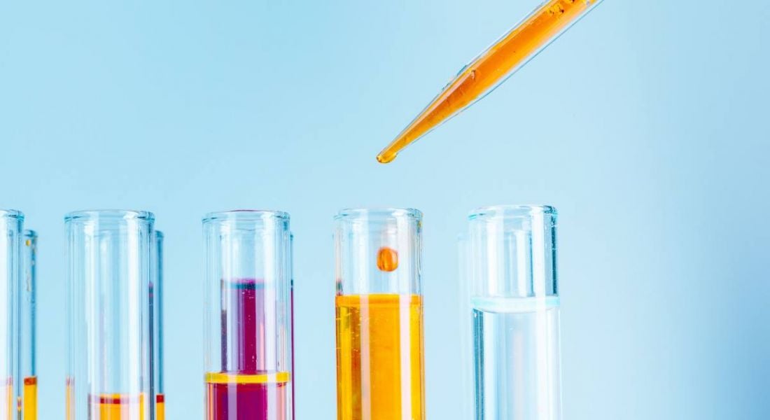 A pipette is adding orange liquid to test tubes against a pale blue background.