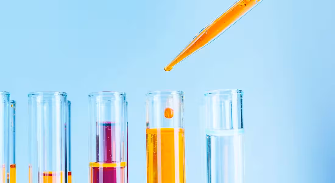 A pipette is adding orange liquid to test tubes against a pale blue background.