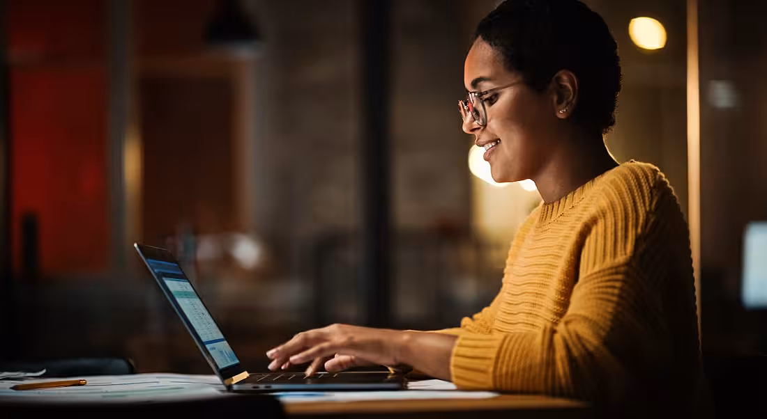 A data professional is working on a laptop while wearing a bright yellow jumper and glasses.