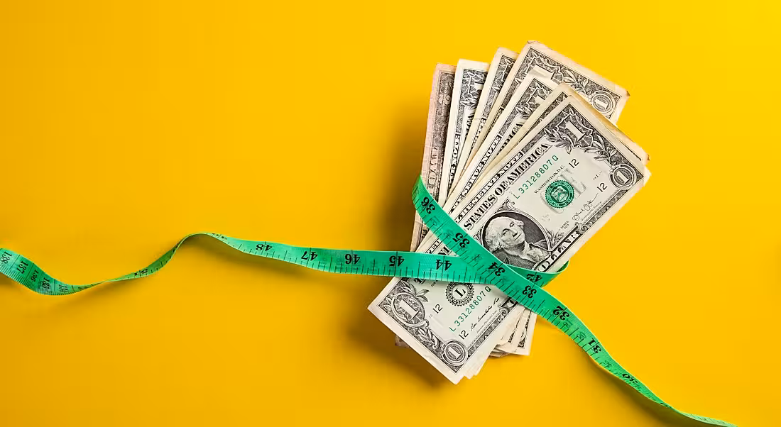 A pile of dollars has a green measuring tape around it on a yellow background.