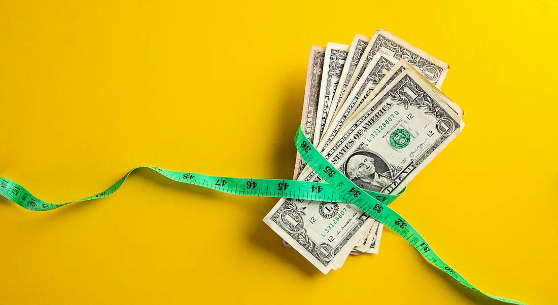 A pile of dollars has a green measuring tape around it on a yellow background.