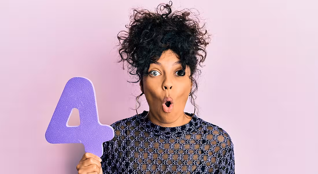 A woman is holding up a purple number four sign and standing against a pink wall.