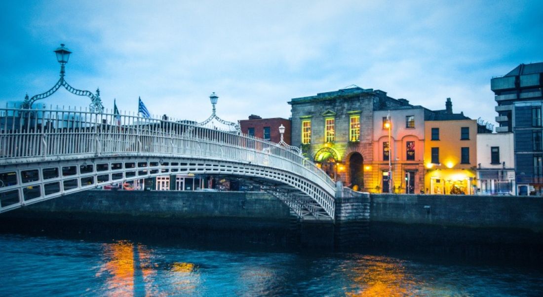 Ha’penny Bridge over the River Liffey in Dublin at dusk. The sky is dark blue and there are lights on buildings in the background.