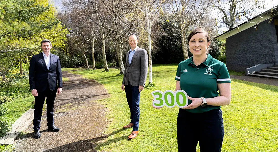 Three people stand socially distanced in a park. The woman in front holds a sign with the number 300 on it.