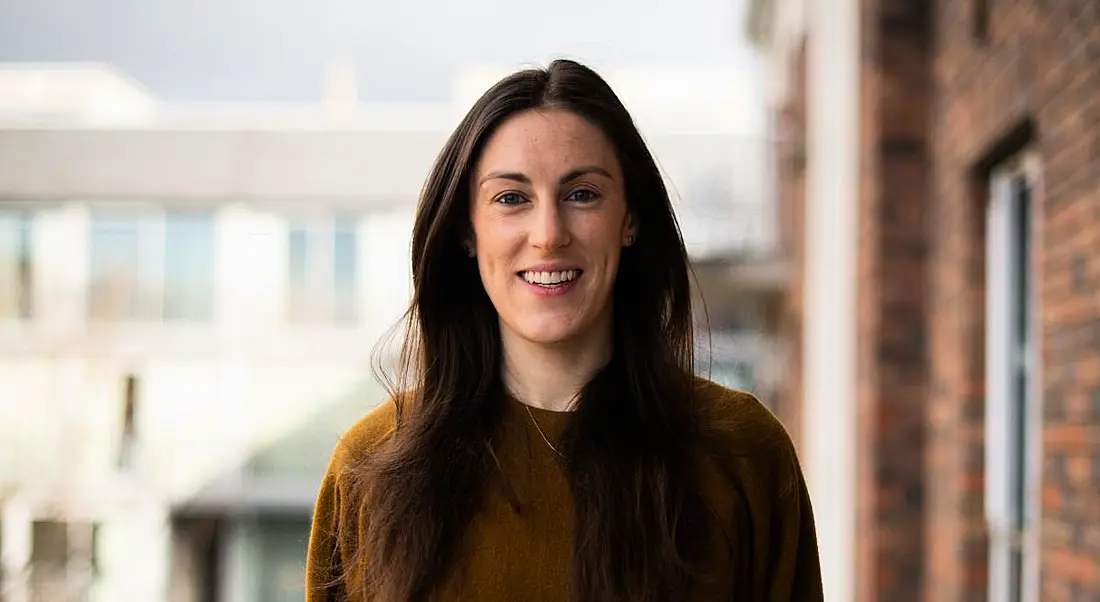 Elaine O’Dwyer, who works at the intersection between data and life sciences, is standing outdoors and wearing a brown jumper while smiling into the camera.