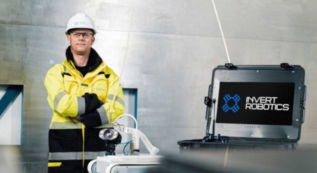 An Invert Robotics employee is standing beside a screen with company branding on it. He is wearing a hard hat and a high-vis jacket.