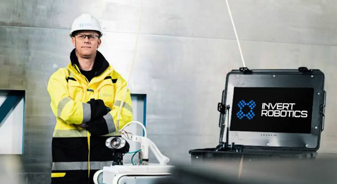 An Invert Robotics employee is standing beside a screen with company branding on it. He is wearing a hard hat and a high-vis jacket.