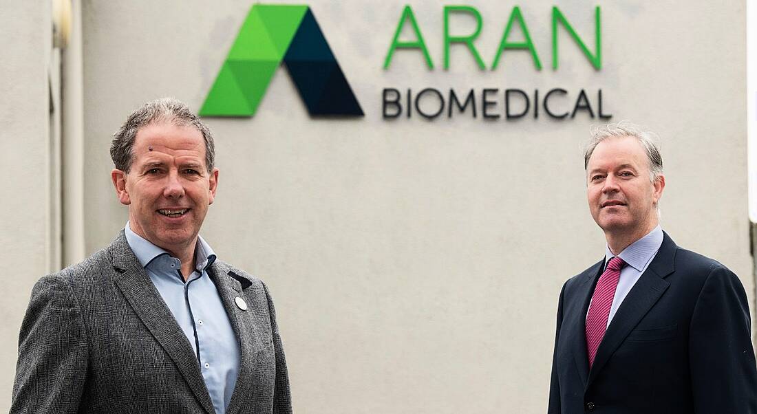 Two men, one in a grey suit and the other in a navy one, stand outside a white building bearing the Aran Biomedical logo.