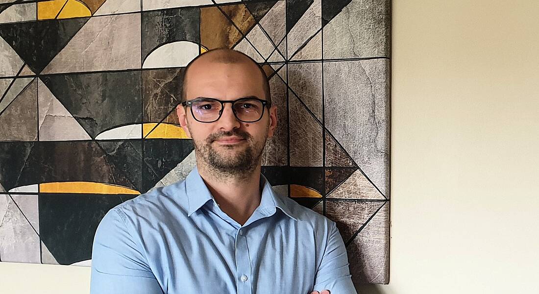 A man in a light blue shirt stands with his arms folded. An abstract painting behind him consists of geometric shapes in earth tones.