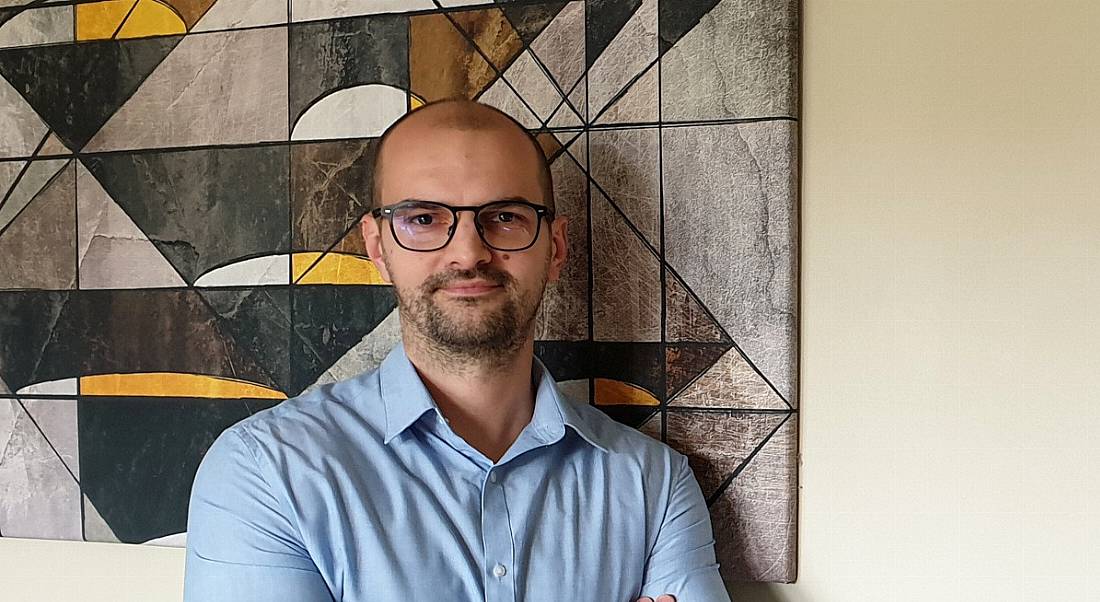 A man in a light blue shirt stands with his arms folded. An abstract painting behind him consists of geometric shapes in earth tones.