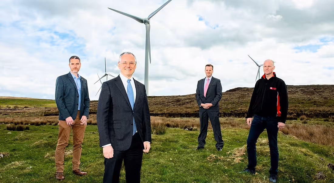 Four men in suits are standing outdoors in front of a wind turbine and looking into the camera.