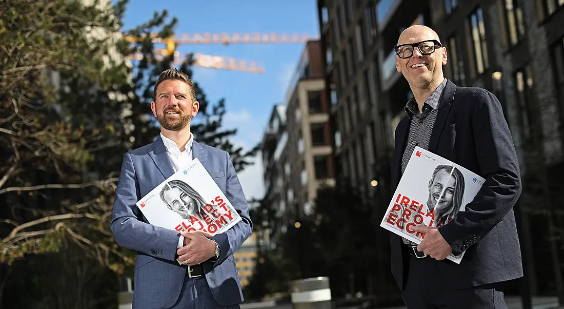 Two men are standing outdoors on a sunny day holding the first edition of an annual report on the project economy in Ireland.