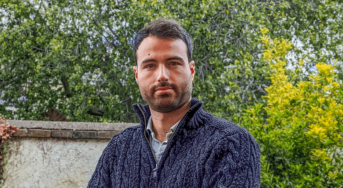 A man in a woollen jumper pictured outside against a background of green leaves, dappled with sunshine.