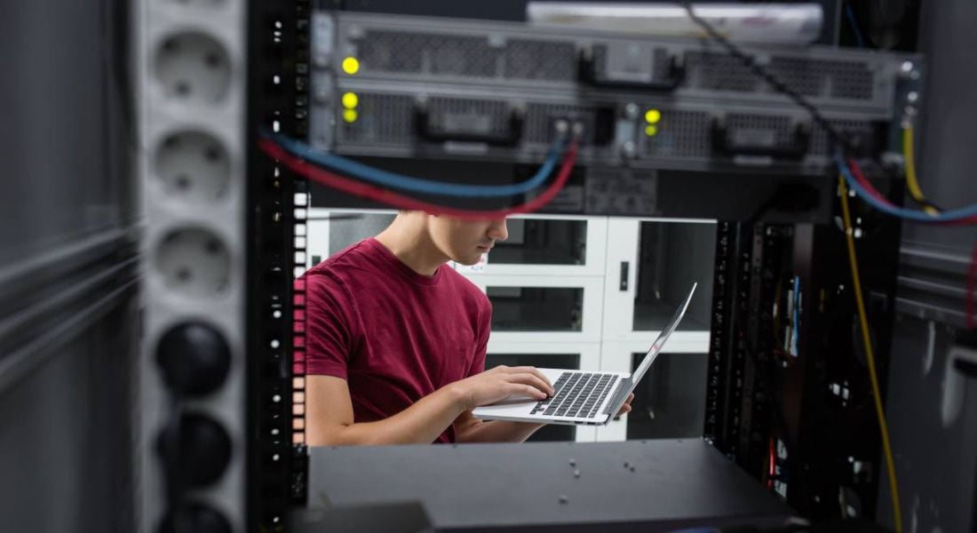 A man in a red T-shirt is standing in a server room and working on a laptop.