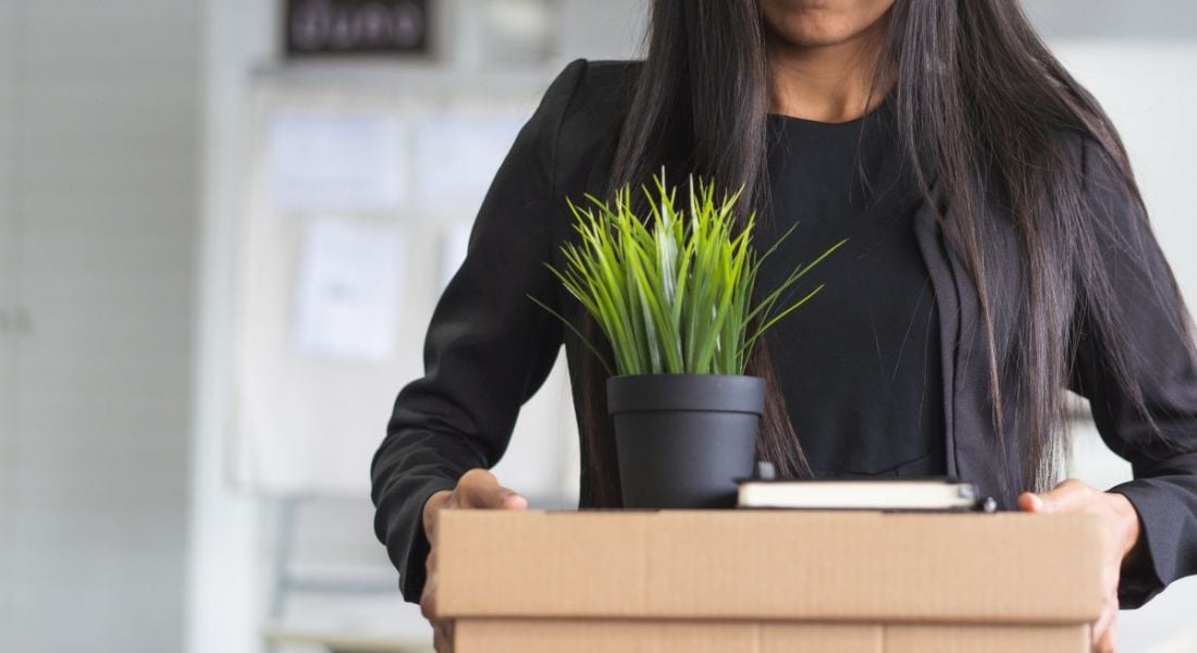A professional woman is carrying a cardboard box of belongings with a plant on top as she quits her job.