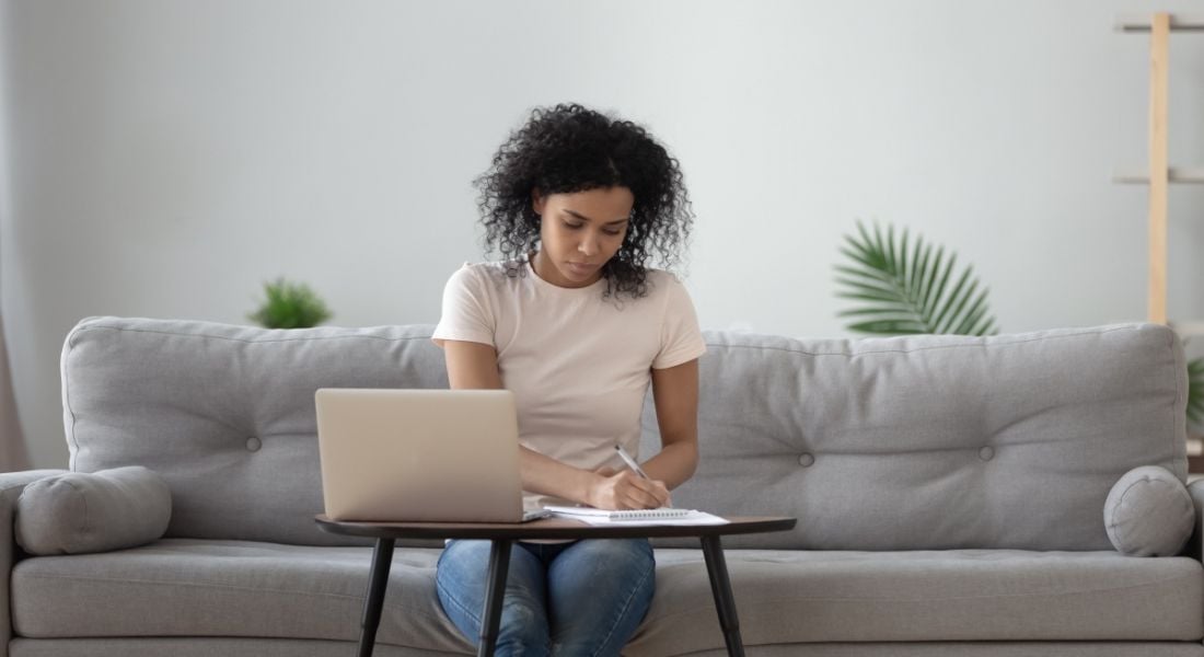 A woman is working remotely on a grey couch at a small desk with a laptop on it.