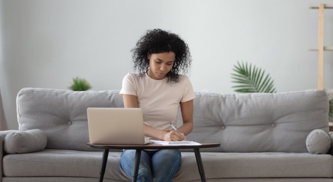 A woman is working remotely on a grey couch at a small desk with a laptop on it.