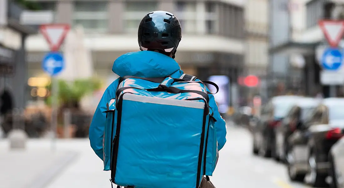 A gig worker is carrying a thermal blue bag on his back, while stopped in the middle of a city street.