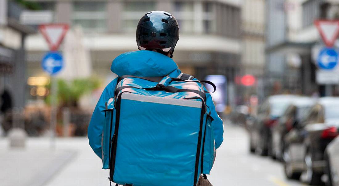 A gig worker is carrying a thermal blue bag on his back, while stopped in the middle of a city street.