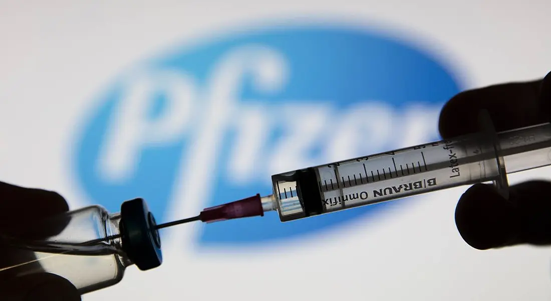 A needle is being inserted into a vaccine vial, with the Pfizer logo in the background.