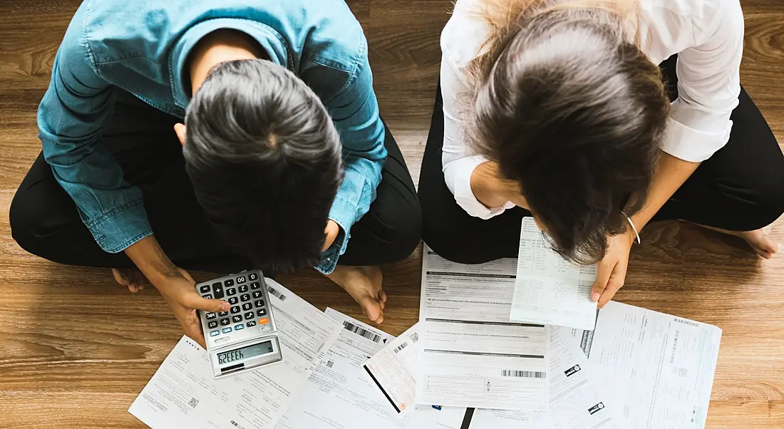 Top view of two young people sitting cross legged on the floor trying to work out tax forms with a calculator.