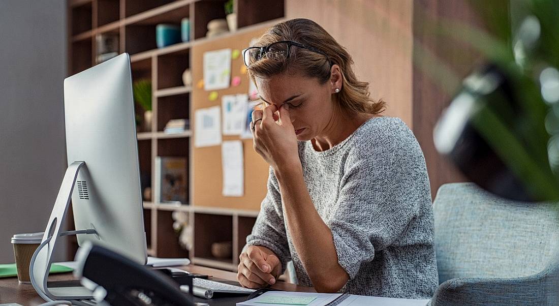 A woman sitting at a desk working at a large computer screen. She is pinching the bridge of her nose, showing she is suffering from burnout.