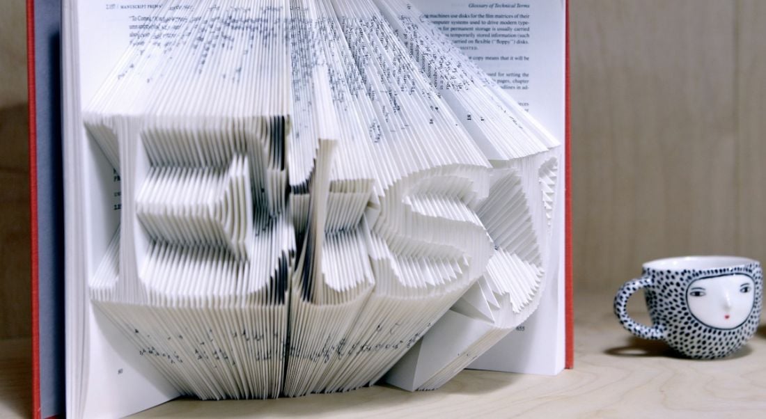 An open book is displayed with pages cut out to spell the word Etsy. It sits on a table beside a small colourful teacup.