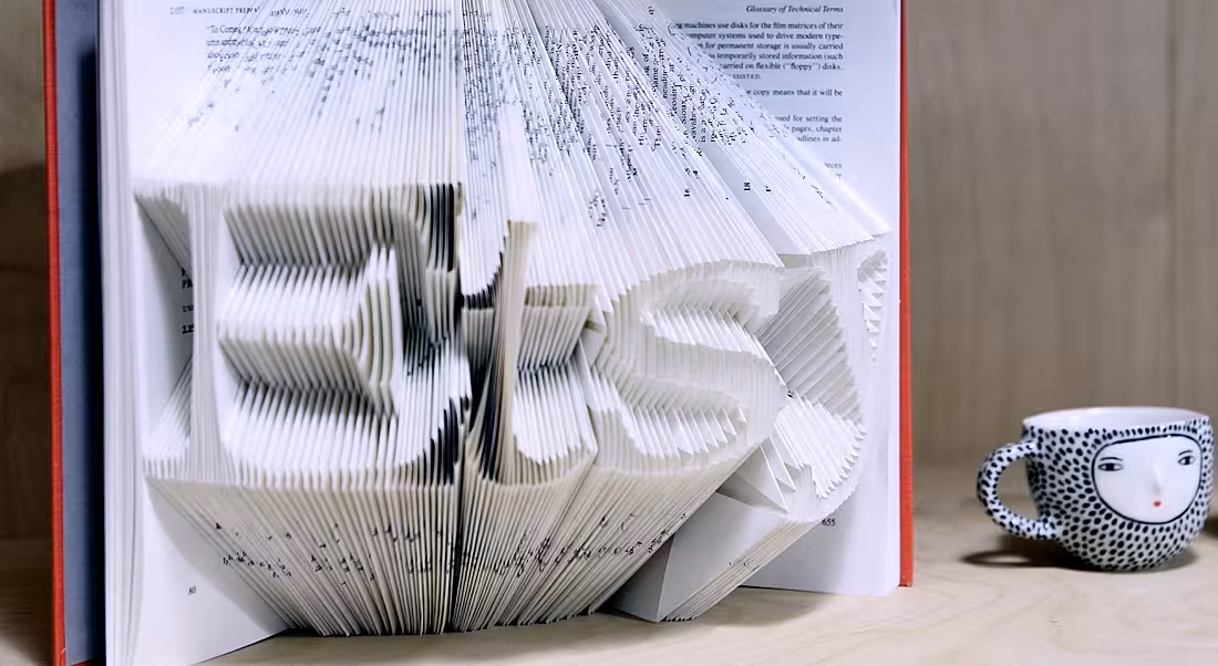 An open book is displayed with pages cut out to spell the word Etsy. It sits on a table beside a small colourful teacup.